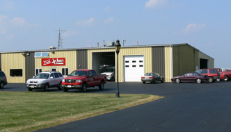 Metal building with garage doors and cars parked out front with the Jim’s Plumbing sign on the side of the building